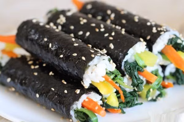 Ngoài ra, cơm cuộn Hàn Quốc (kimbap) cũng dễ làm tại nhà. Trải rong biển, dàn cơm đều, xếp trứng tráng mỏng, cà rốt, dưa chuột và xúc xích lên trên, cuộn chặt tay, cắt khoanh và rắc mè rang, bữa ăn vừa ngon vừa tiện lợi để mang đi. Ảnh minh họa