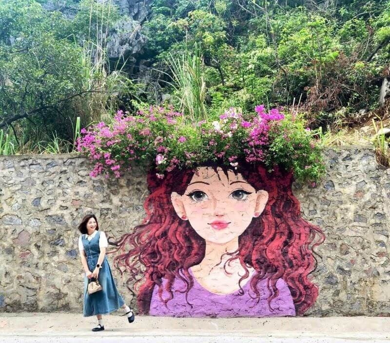 Mỗi bức tranh là một phong cách riêng: có cô gái dịu dàng trong tà áo dài, người hiện đại với mái tóc xoăn dài, ánh mắt sâu lắng, hay thiếu nữ ngây thơ má ửng hồng. Điểm đặc biệt là tranh luôn gắn với những chậu hoa giấy rực rỡ – đôi khi trở thành mái tóc, chiếc mũ hay vương miện tự nhiên, tăng thêm vẻ mềm mại và lãng mạn. Ảnh Huyền Khánh Vũ