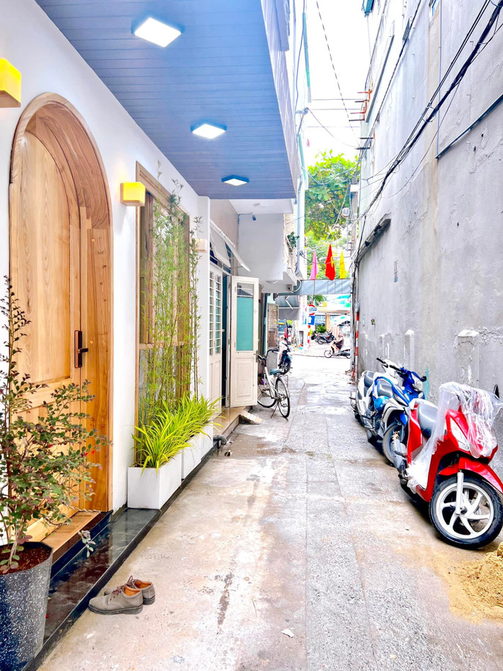 Ngôi nhà ống nằm ở trong con hẻm với mặt tiền đẹp. Gia chủ đã thiết kế một cửa gỗ mái vòm và cửa kính lớn, bổ sung thêm vài chậu cây bằng bê-tông dẻo sơn trắng cho sinh động. Chị lựa chọn trồng trúc và cây cảnh loại lá kim. Trúc có ý nghĩa là "Phúc" - thể hiện ước mong về cuộc sống hạnh phúc, tài lộc dồi dào.