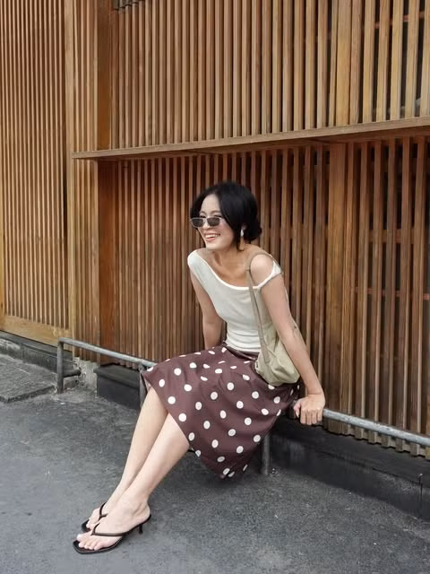 Áo trắng lệch vai, chân váy chấm bi và dép xỏ ngón mang đến vẻ thoải mái. Kính râm dáng nhỏ, túi vải và tóc búi thấp gợi phong cách retro, phù hợp khi dạo phố.