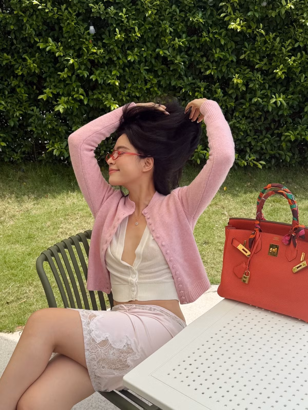 Ở phong cách đời thường, cô mặc cardigan hồng bên ngoài áo trắng dáng ngắn, kết hợp chân váy ren. Kính gọng đỏ và túi cùng tông tạo nét trẻ trung cho bộ đồ.