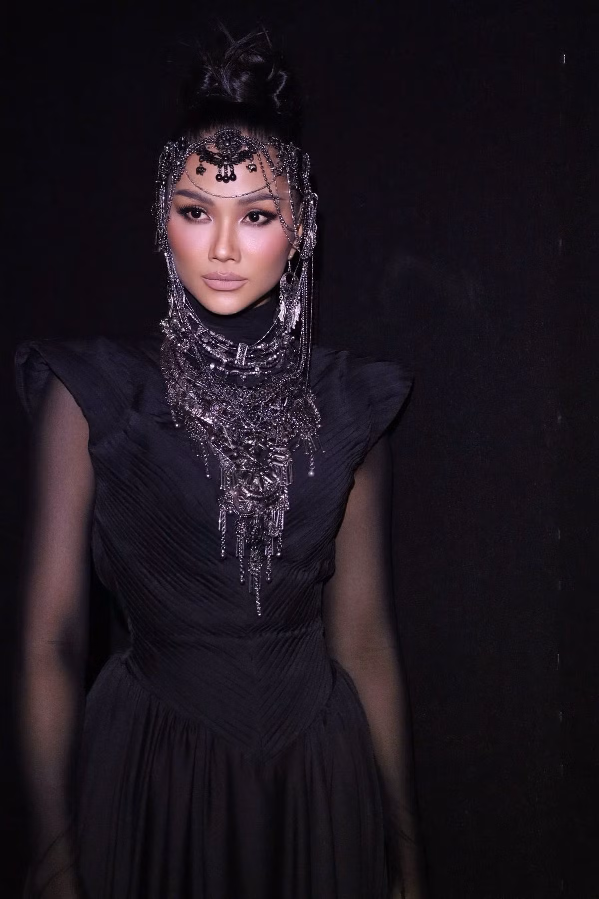 Trên thảm đỏ, H’hen Niê khoác lên mình bộ trang phục đen dáng dài, thiết kế ôm gọn phần thân trên với chi tiết vai dựng sắc nét kết hợp tay áo xuyên thấu, tạo cảm giác vừa cá tính vừa bí ẩn. Phần chân váy rủ mềm, chuyển động uyển chuyển theo từng bước đi, giúp tôn lên vóc dáng săn chắc và cân đối.