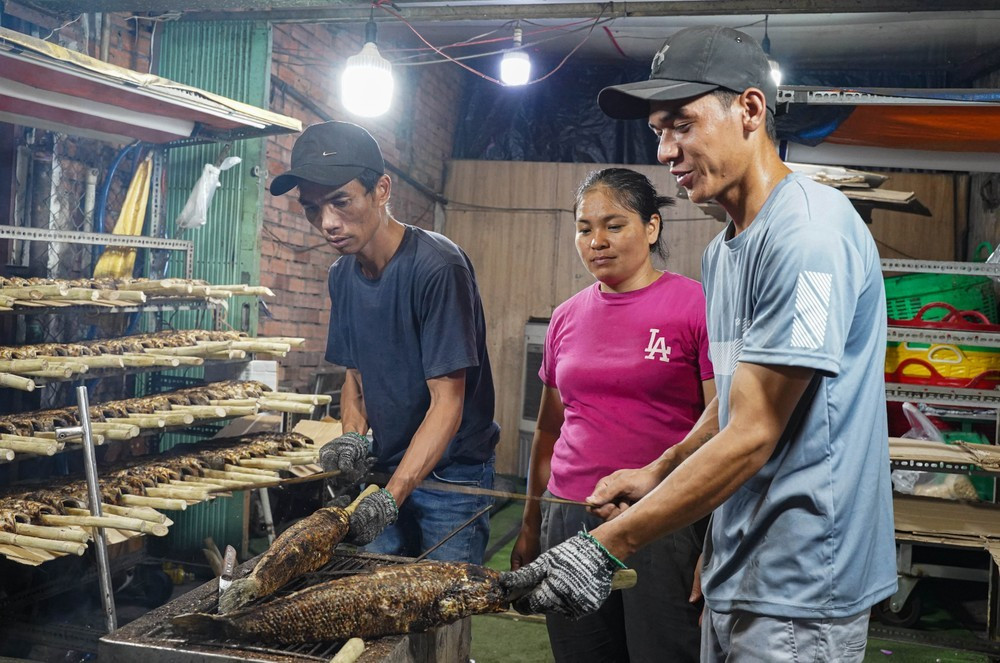 Anh Danh Khá (bìa phải, quê Bạc Liêu) lần đầu tiên lên phụ gia đình anh Văn Phú (bìa trái, chủ tiệm) nướng cá lóc ngày cao điểm. "Năm nay, tính đến thời điểm hiện tại thấy sức mua của người dân có phần giảm, độ tất bật, nhộn nhịp của tuyến đường này trong đêm trước ngày vía Thần Tài không bằng năm rồi. Nhưng tiệm của vợ chồng tôi vẫn chuẩn bị 500-600 con cá cho ngày mai" - Anh Văn Phú chia sẻ.