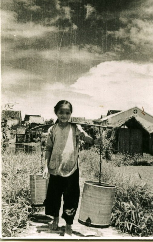 Bé gái gánh nước bằng hai thùng sắt tây cỡ lớn.