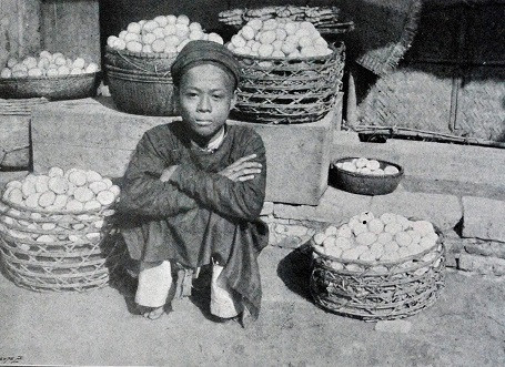 Một cậu bé ngồi bán những giỏ đựng đầy trứng tại một phiên chợ xưa.