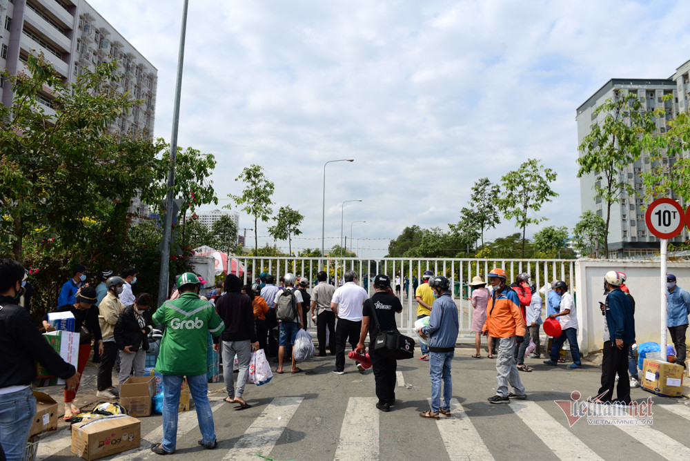 Các lực lượng trong đội bảo vệ khu cách ly liên lục phát loa, người dân chỉ nên mang khẩu trang, thuốc, đồ dùng phụ nữ đến cho người thân. Dù vậy, lượng người mang thau, chăn màn, nệm, móc quần áo, đồ chà nhà vệ sinh… đến tiếp tế cho người cách ly vẫn mỗi lúc một đông. Họ đứng ở khắp cổng ra vào, tràn ra cả đường đi mặc trời Sài Gòn nắng chang chang. (Nhiều người vẫn đội nắng mang đồ đến cho người thân bị cách ly)