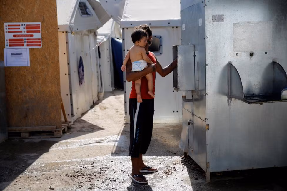 Một người tị nạn Syria vừa bế con vừa lấy nước vào chai trong trại tị nạn ở Chios, Hy Lạp, hôm 7/9.