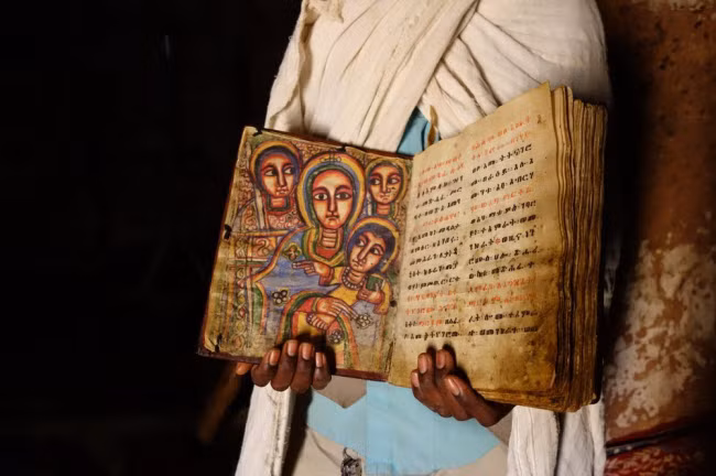 Một linh mục cầm cuốn kinh thánh làm bằng da dê, được minh họa bằng tranh vẽ tay và chữ nghệ thuật.