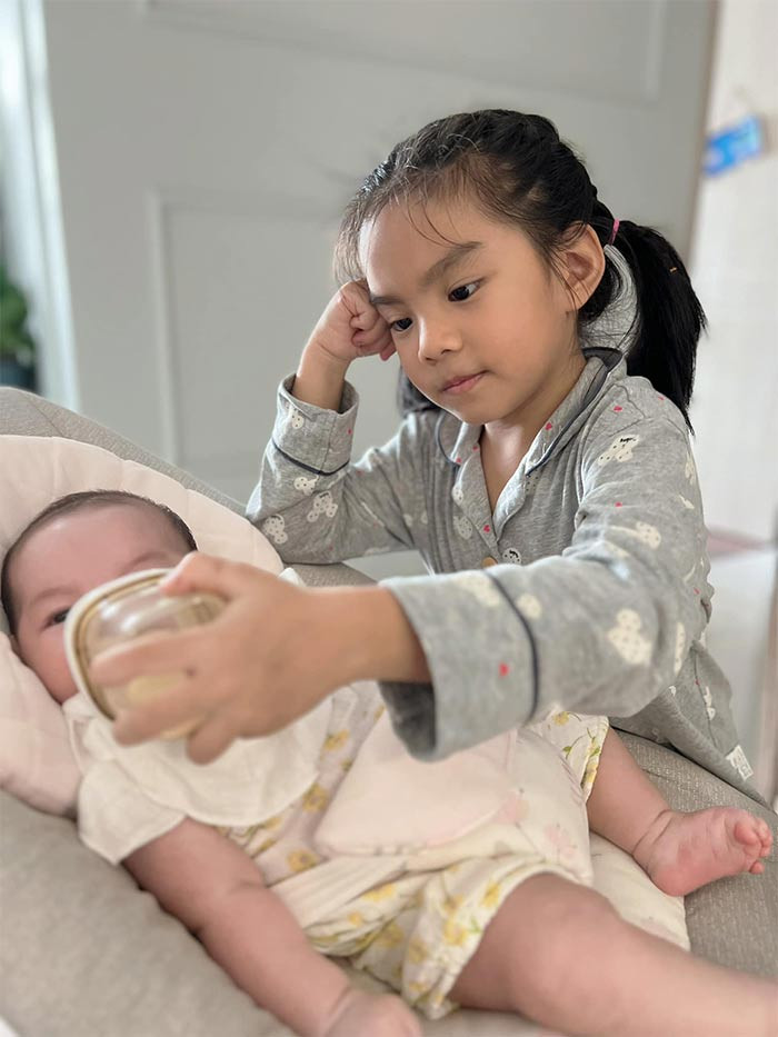 Chị gái thứ hai giúp mẹ cho em uống sữa.