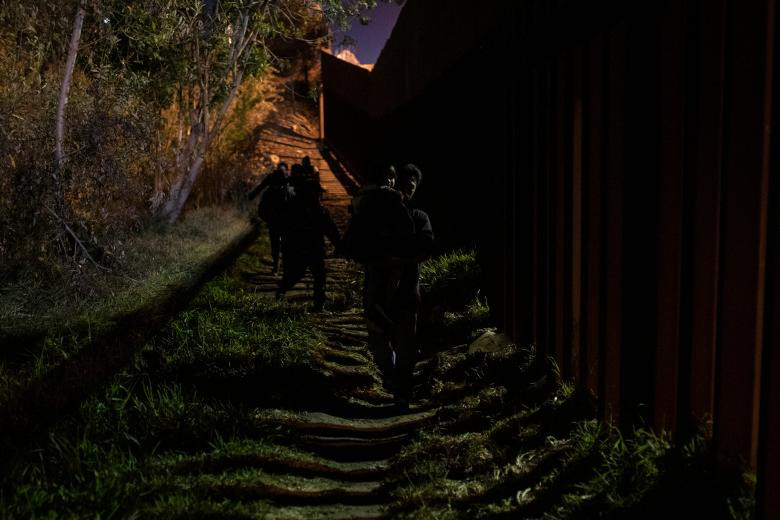 Những người dân di cư đi bộ cạnh bức tường biên giới trước khi vượt biên từ Mexico vào Mỹ lúc trời tối hôm 3/12.