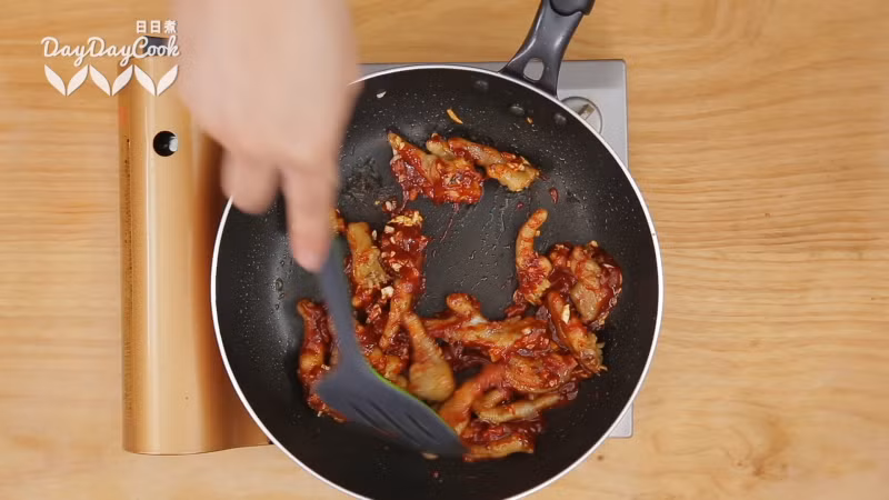 Cho một ít dầu vào chảo phi tỏi cho thơm rồi cho chân gà vào đảo. Tiếp theo cho sốt ớt vào và bật to lửa. Thêm khoảng 200ml nước vào trộn đều rồi tiếp tục nấu đến khi nước sốt sánh lại.