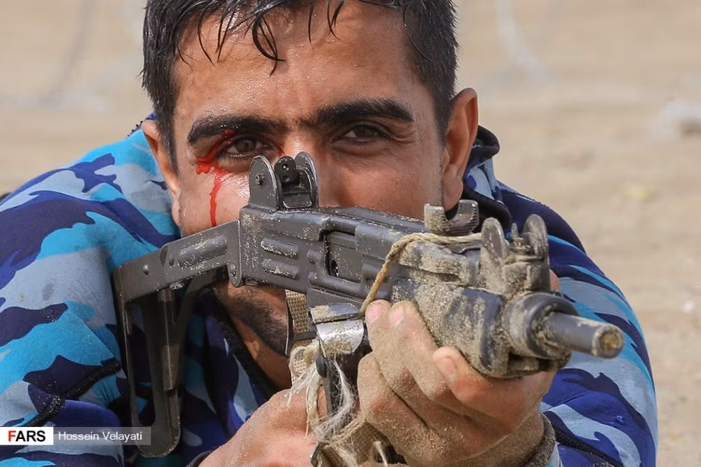 Một linh hải quân Iran bị thương chảy máu mắt trong cuộc đổ bộ, khẩu tiểu liên Uzi mà người lính này mang theo cũng bị cát bịt kín miệng súng. Nguồn ảnh: Sina.
