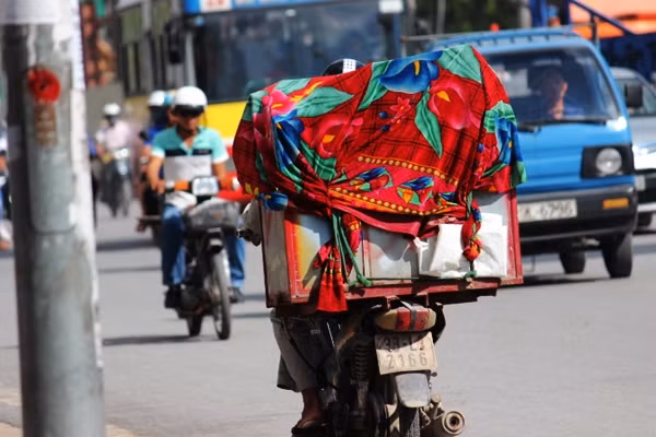 Một chiếc xe máy cũ, xuống cấp chở theo thùng đá viên, bên ngoài được phủ lớp chăn kỳ dị vô tư đi ngược chiều trên đường Cầu Diễn.