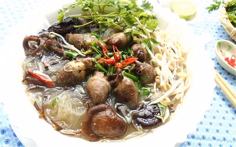 Đun sôi 300ml nước dùng gà, cho miến vào, trụng sơ qua cho sợi miến mềm, vớt ra rổ. Cho miến vào tô, chan nước dùng gà vào. Thêm hỗn hợp tim gà, hành lá, ngò rí, rau răm, giá đỗ, ớt cắt nhỏ lên trên và thưởng thức. Ảnh: Cooky.vn.