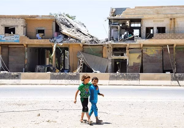 Hai em nhỏ khoác vai nhau đi qua những tòa nhà đổ nát ở Aleppo. Ảnh: FNA.