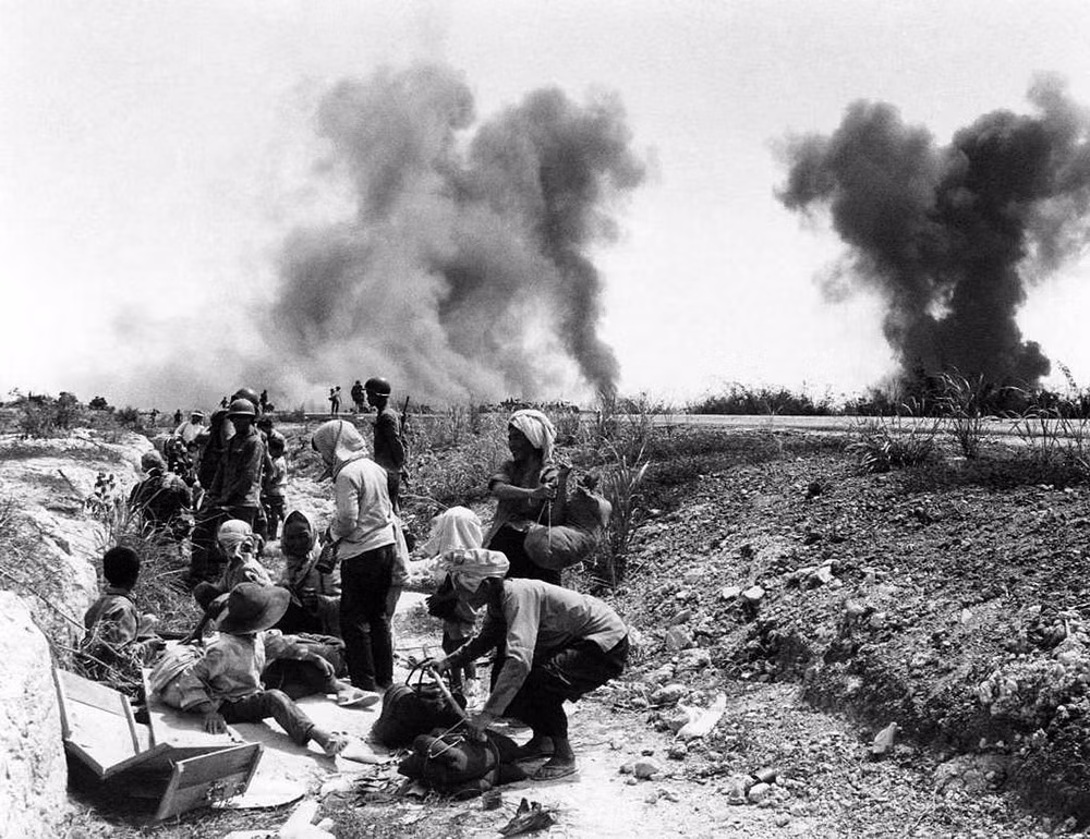 Người dân di tản từ thị xã An Lộc nghỉ ngơi ở một mương cạn ven đường, tháng 2/1972. Thị xã An Lộc nằm cách đó 8 dặm về phía Bắc, đang bị quân Giải phóng bao vây. Những cột khói bốc lên từ hoạt động không kích để giải vây cho thị xã của Mỹ và quân đội Sài Gòn. Ảnh: AP Photo/ Nick Ut