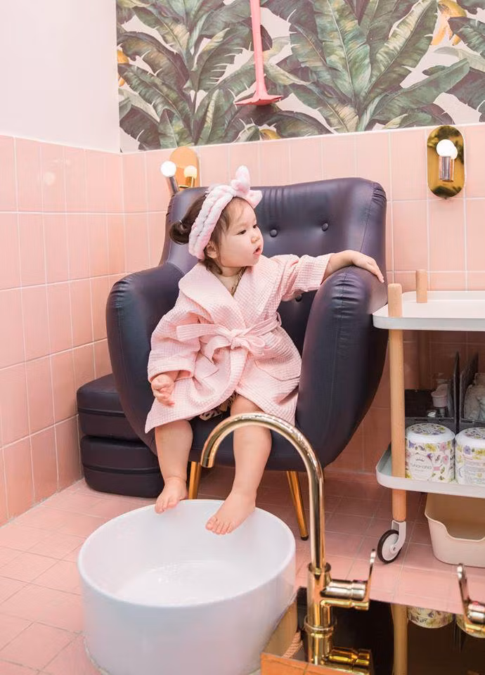Bé Myla vận bộ trang phục màu hồng, quấn chiếc nơ hồng trên đầu ngồi thư giãn trên ghế để nhân viên phục vụ massage chân thư giãn.