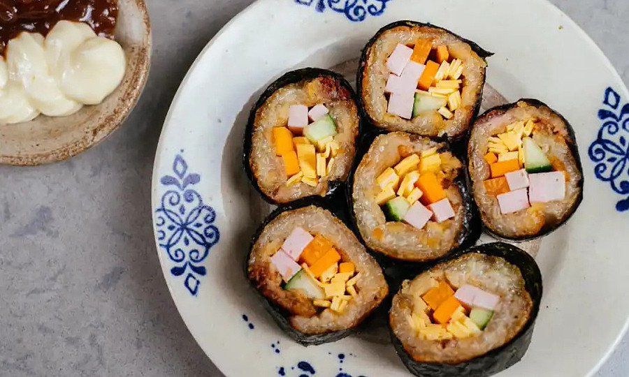 Để thực hiện món ăn, cần cắt mỏng bánh chưng, dầm nhuyễn rồi rán bằng nước. Khi bánh chưng se mặt thì tắt bếp, không nên rán vàng giòn sẽ khó cuộn. Đặt tấm rong biển lên mặt phẳng, dàn đều bánh chưng tạo độ kết dính rồi đặt các nhân có sẵn hoặc còn tồn đọng sau Tết như giò, xúc xích thái sợi dài, rau củ, trứng rán... vào và cuộn lại thật chặt tay. Dùng dao sắc nhúng nước nóng hoặc thoa chút dầu ăn rồi cắt kimbap thành các miếng vừa ăn. Thưởng thức chấm cùng sốt mayonnaise pha lẫn tương ớt, tương cà tùy khẩu vị. Ảnh Internet