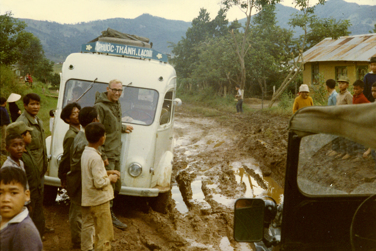 Ông Tom Petersen tươi cười bên chiếc xe khách đang bị mắc kẹt tại một đoạn đường ngập bùn trên đường từ Đà Lạt về căn cứ của mình 9 (quốc lộ 20). Ảnh: Tom Petersen.
