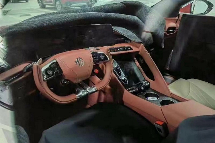 Vô lăng này được bọc da màu nâu, tích hợp phím chức năng và 2 lẫy chuyển số thể thao. Trên vô lăng còn có nút màu đỏ để khởi động máy và nút màu đen để chọn chế độ lái. Phía sau vô lăng là màn hình cong khá lớn, đặt nổi trên mặt táp-lô. Trên cụm điều khiển trung tâm của xe có vẻ còn có thêm một màn hình cảm ứng nằm dọc nữa.