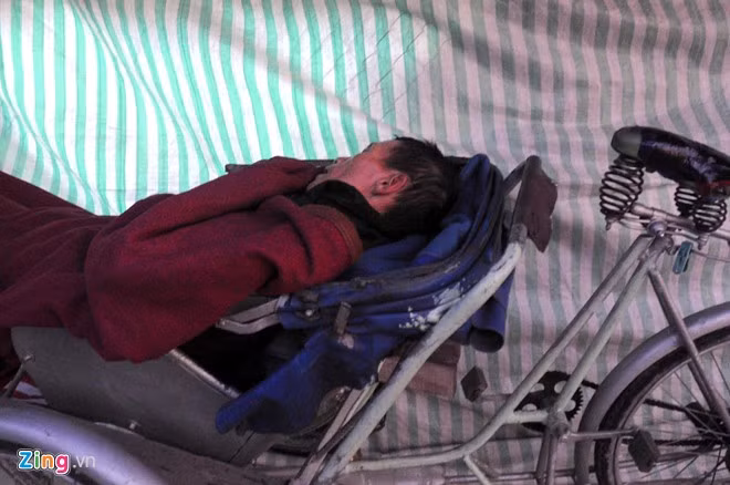 Người đàn ông đạp xích lô tránh cái giá lạnh buổi sáng sau tấm vải, ông cố ngủ thêm một lúc trong khi đường phố đã đông người.