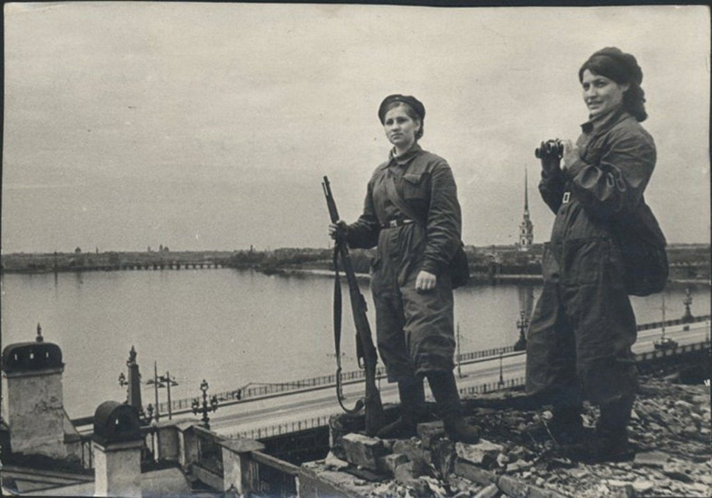 Trong khi đó, một số phụ nữ Liên Xô cầm súng và ống nhòm canh gác bầu trời Leningrad để kịp thời phát hiện máy bay Đức quốc xã.