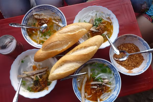 Bánh canh chờ tại Đà Nẵng. Không chỉ như tên gọi là phải “chờ” mới được ăn, khi bạn đến đây phải sẵn sàng tầm lý “tự phục vụ” nếu không muốn mang cái bụng đói ra về. Tuy vậy, có lẽ chẳng ai thấy phiền lắm vì bánh canh chờ “vừa ngon vừa rẻ”. Quán ở cuối đường Trần Hưng Đạo, bàn ghế đơn sơ được đặt trên dải đất trống, muốn ăn cũng phải tự phục vụ từ tô cháo đến bánh mì và giá chỉ 7.000đ/ tô. (Ảnh: dulichdanang.vn).