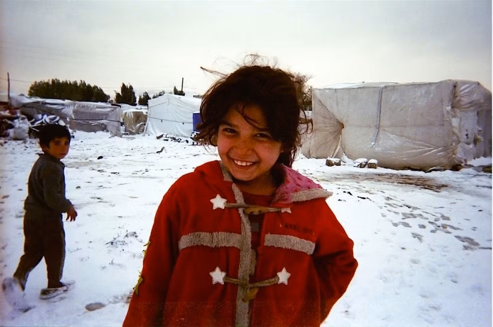 Một bé gái tị nạn mặc chiếc áo khoác đỏ sờn cũ, tươi cười trong khu trại phủ đầy tuyết trắng.