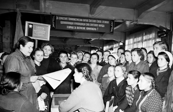 Các nữ công nhân tại một nhà máy lắp ráp đồng hồ đang chăm chú lắng nghe một người đọc báo.