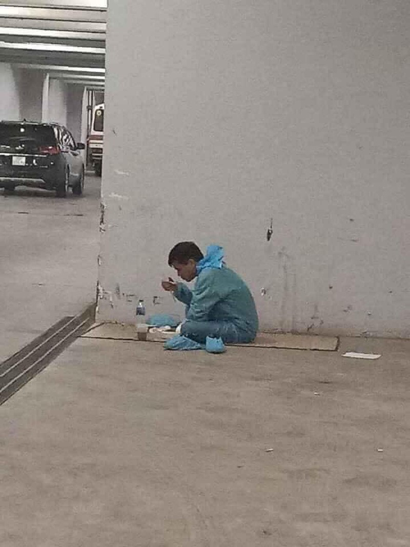 Tài xế lái xe bệnh nhân cách ly ăn vội suất cơm hộp trong góc hầm để xe. Anh vẫn mặc nguyên đồ bảo hộ, trải tạm manh chiếu nhỏ để có chỗ ngồi dưới nền bê tông bụi bặm.