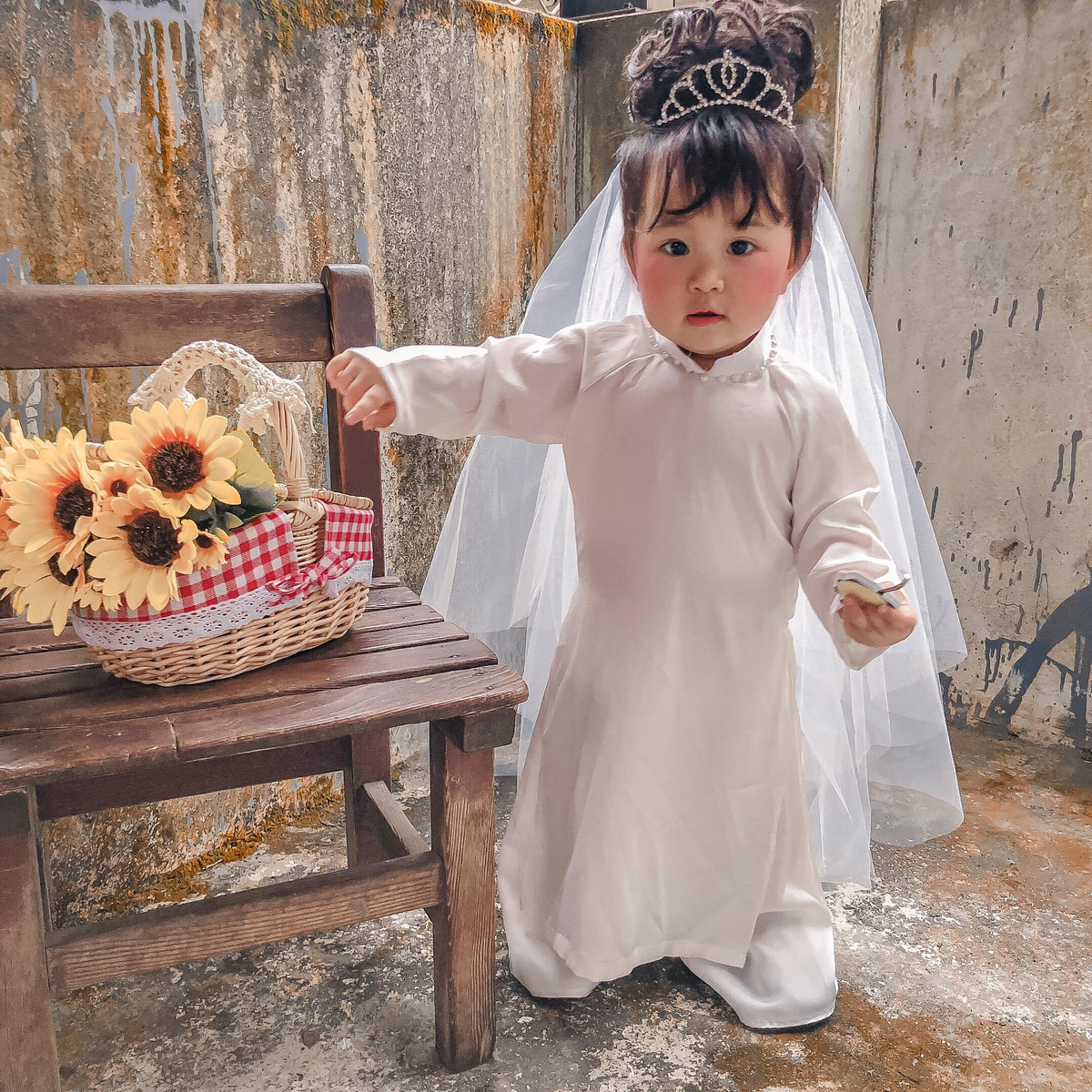 Trong ảnh, em bé cosplay cô dâu mini mặc một chiếc áo dài trắng tinh khôi, đeo vương miện xinh xinh và thêm chiếc khăn voan đội đầu. Bối cảnh của bộ ảnh cũng vô cùng đơn giản, chỉ là ngõ nhỏ với bức tường rêu phong, giỏ hoa và chiếc ghế gỗ, thế là có những tấm hình cưới phong cách cũ.