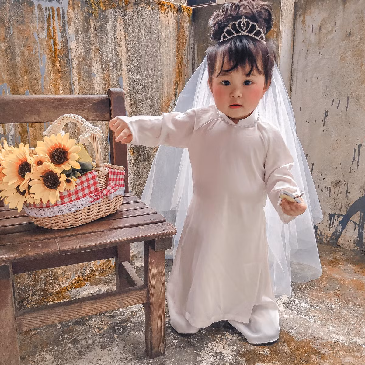 Trong ảnh, em bé cosplay cô dâu mini mặc một chiếc áo dài trắng tinh khôi, đeo vương miện xinh xinh và thêm chiếc khăn voan đội đầu. Bối cảnh của bộ ảnh cũng vô cùng đơn giản, chỉ là ngõ nhỏ với bức tường rêu phong, giỏ hoa và chiếc ghế gỗ, thế là có những tấm hình cưới phong cách cũ.