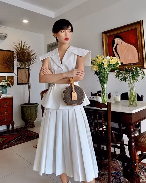 Ở nhà cũng phải đẹp là tôn chỉ của Khánh Linh. Với áo peplum và chân váy tiệp màu trắng, cô nàng chọn túi Boite Chapeau Souple của Louis Vuitton làm phụ kiện chấm phá, vừa thanh lịch vừa thời thượng.