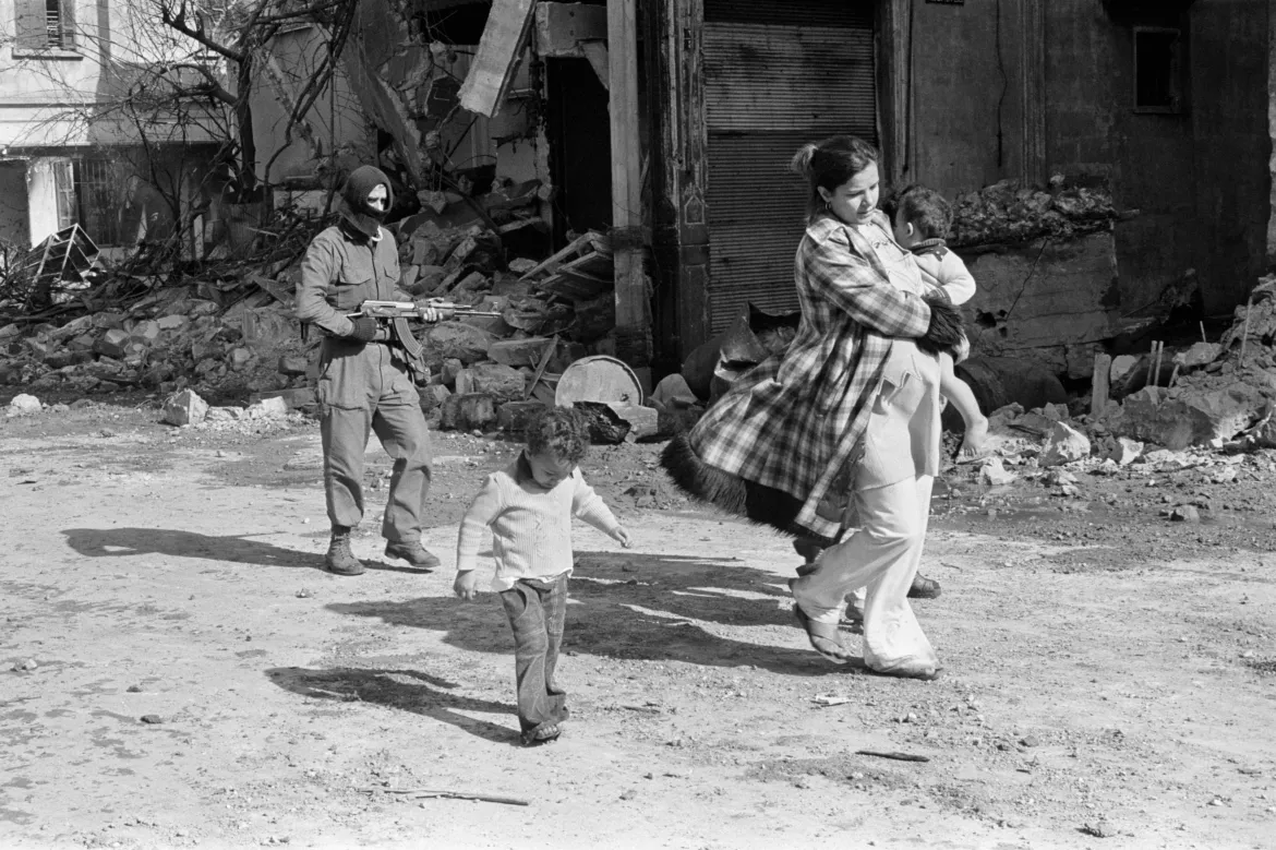 Người phụ nữ bế con nhỏ đi trên con phố ở Karantina, Beirut, ngày 19/8/1976. Phía sau cô là một chiến binh mang theo súng.