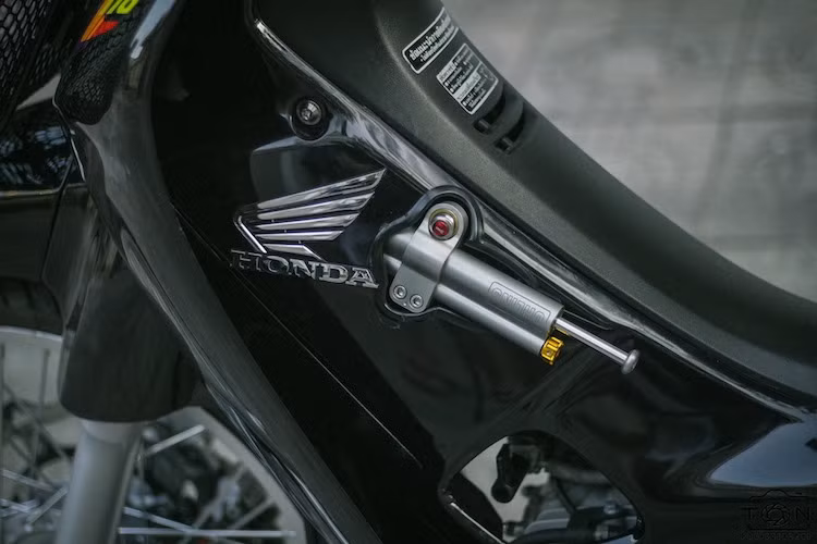 Trong khi đó, gắn ở chảng ba dưới là cây trợ lực Ohlins, khiến phần đầu của chiếc Wave ổn định hơn khi chạy tốc độ cao. Xe đi tem nguyên bản của Wave 110 Thái Lan trên nền sơn đen, với điểm nhấn là logo Honda cánh chim mạ chrome sang trọng hai bên yếm và một số vị trí bắt ốc titan Proti.