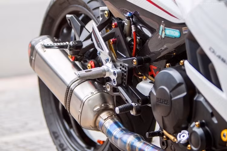 Với cây pô Akrapovic cùng cổ pô bằng titan gia nhiệt ánh xanh đẹp mắt, tiếng nổ từ động cơ của Exciter 150 đã trở nên trầm ấm hơn. Nằm ngay cạnh cây pô là dàn để chân Rizoma bằng nhôm với khả năng điều chỉnh vị trí tự do.