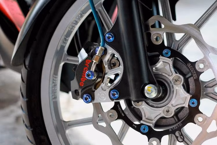 Ở phía trước, hệ thống phanh đĩa nguyên bản của chiếc xe đã được thay thế bằng loạt phụ kiện "hàng hiệu" gồm đĩa Galfer và heo Brembo Billet 2 pis đối xứng, bắt vào phuộc xe nhờ pad nhôm CNC sắc sảo. Những con ốc Proti giúp hệ thống phanh trước của xe càng trở nên nổi bật hơn.