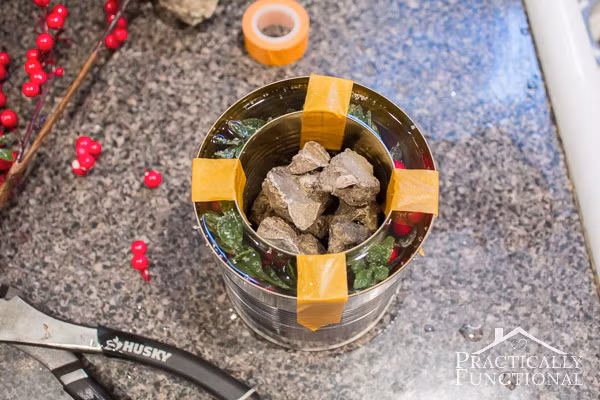 Sau đó, bạn cắt những nhành cây xanh và quả mọng thả vào khe giữa hai lon. Bạn cần chú ý dàn đều những nhành cây và quả ra để chiếc đèn trông đẹp mắt.