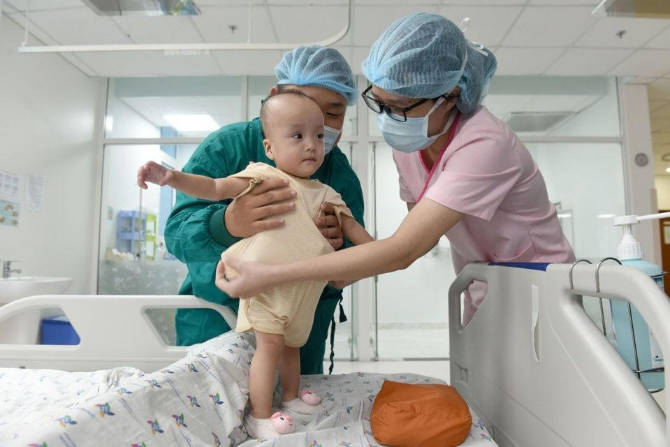 Giờ đây, đôi chân của hai bé khép sát nhau, cứng cáp hơn, đã có thể vịn đứng thời gian ngắn khi có sự hỗ trợ của ba mẹ hoặc nhân viên y tế.