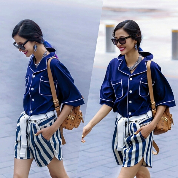 Hoàng Thuỳ chọn một thiết kế sơ mi đậm chất pajamas với nền xanh tím than và viền trắng nổi bật, kết hợp sur ton cùng quần shorts kẻ xanh-trắng to bản.