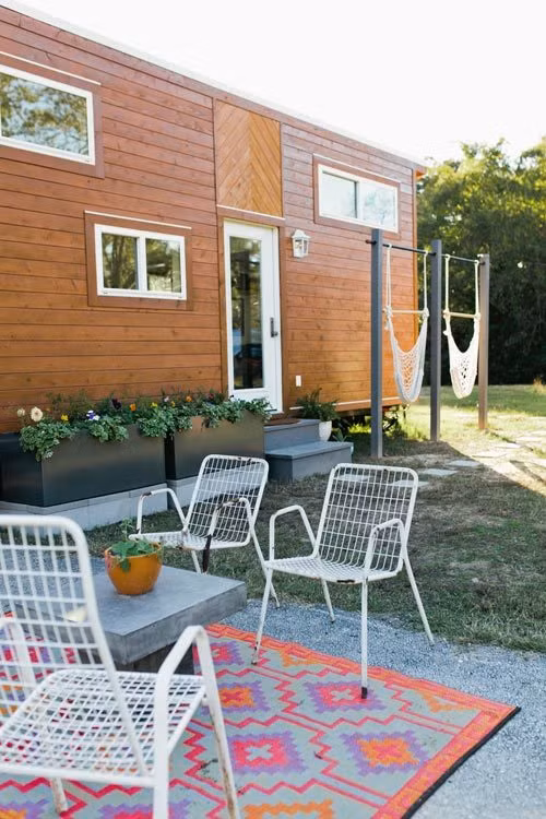 Bên ngoài căn nhà được trang trí bởi những họa tiết đơn giản từ chất liệu gỗ. Những mảng ghép hình chữ V cũng được bố trí tại một số điểm bên ngoài, tạo điểm nhấn cho phần ngoại thất. Cùng với đó, đôi vợ chồng trẻ cũng bố trí một bộ bàn ghế, hai chiếc xích đu để phục vụ nhu cầu giải trí, nghỉ ngơi ngoài trời. Quanh nhà, Kelly khéo léo trồng những khóm hoa, khiến tổng thể bên ngoài của ngôi nhà thêm phần tươi tắn, sống động.