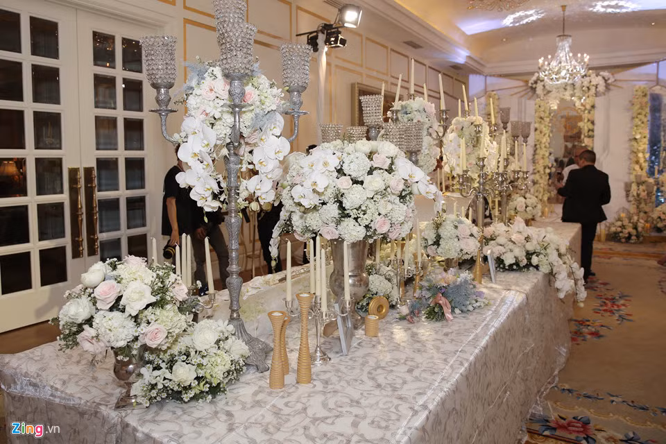 Không gian tiệc cưới được trang trí cầu kì, lộng lẫy với tông màu trắng chủ đạo. Trên đường vào sảnh tiệc là dãy bàn dài gồm nhiều tháp nến, hoa tươi và ảnh kỉ niệm của hai vợ chồng. Những loại hoa như hoa hồng, cẩm tú cầu và địa lan được dùng để trang trí.