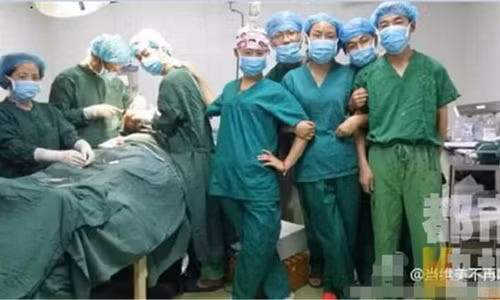 Ba bác sĩ đang khâu vết thương cho bệnh nhân, trong khi những người khác đứng tạo dáng để chụp ảnh ngay bên bàn mổ.