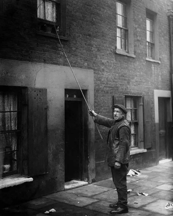 Một người làm nghề "Knocker-Up" ở London, năm 1929. Trước khi có đồng hồ báo thức, có những người sẽ được trả tiền để đánh thức khách hàng của họ đi làm, bằng cách gõ vào cửa hay cửa sổ nhà họ bằng một cây gậy.