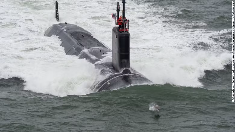 Cá heo bơi trước mũi tàu ngầm USS John Warner (SSN-785) đang rẽ sóng. Đây là tàu ngầm tấn công nhanh chạy nhiên liệu hạt nhân thuộc lớp Virginia. Chúng dài 115 m, rộng 10 m và có tải trọng choán nước 7.800 tấn. Lò phản ứng S9G cho phép tàu di chuyển với vận tốc tôi đa đạt 65 km/h và hoạt động liên tục trong khoảng 20 năm mà không cần tiếp nhiên liệu. Tàu được trang bị tên lửa hành trình Tomahawk để tấn công các mục tiêu trong đất liền.