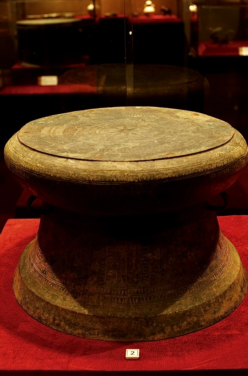 Trống đồng, mặt trang trí hình mặt trời 10 cánh, xung quanh hình chim hạc. Tang trống trang trí thuyền người, thân trống trang trí người nhảy múa, niên đại thế kỷ 5-3 TCN.