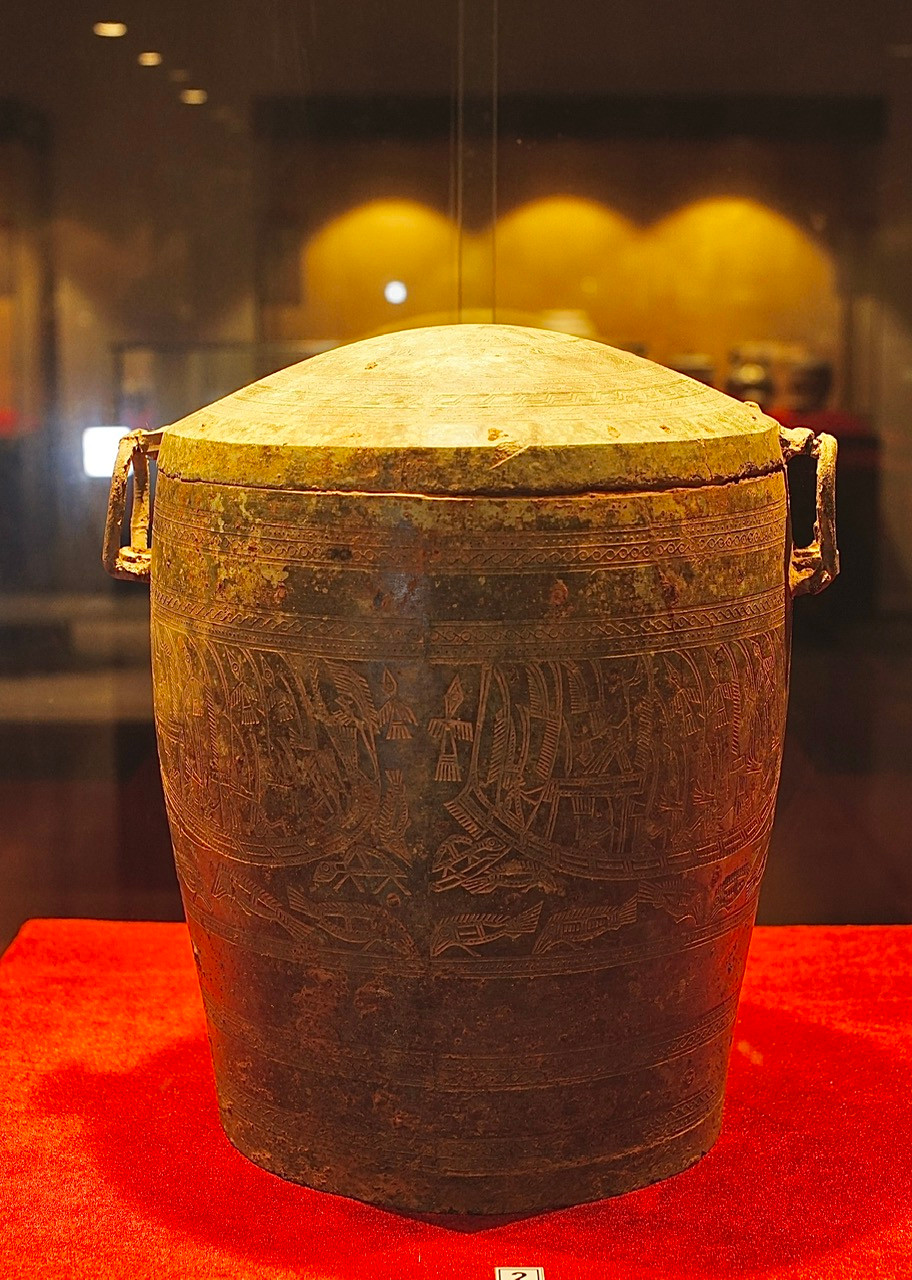 Thạp đồng có nắp, trên nắp là hình mặt trời 10 cánh, thân thạp trang trí thuyền và người đội mũ lông chim, dưới thuyền là hoa văn con cá. Niên đại Thế kỷ 5-3 TCN.