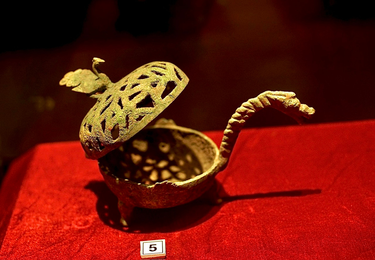 Đèn dầu có nắp bằng đồng, đỉnh nắp trổ thủng tượng phượng, tay cầm hình rồng. Niên đại thế kỷ 1-3.