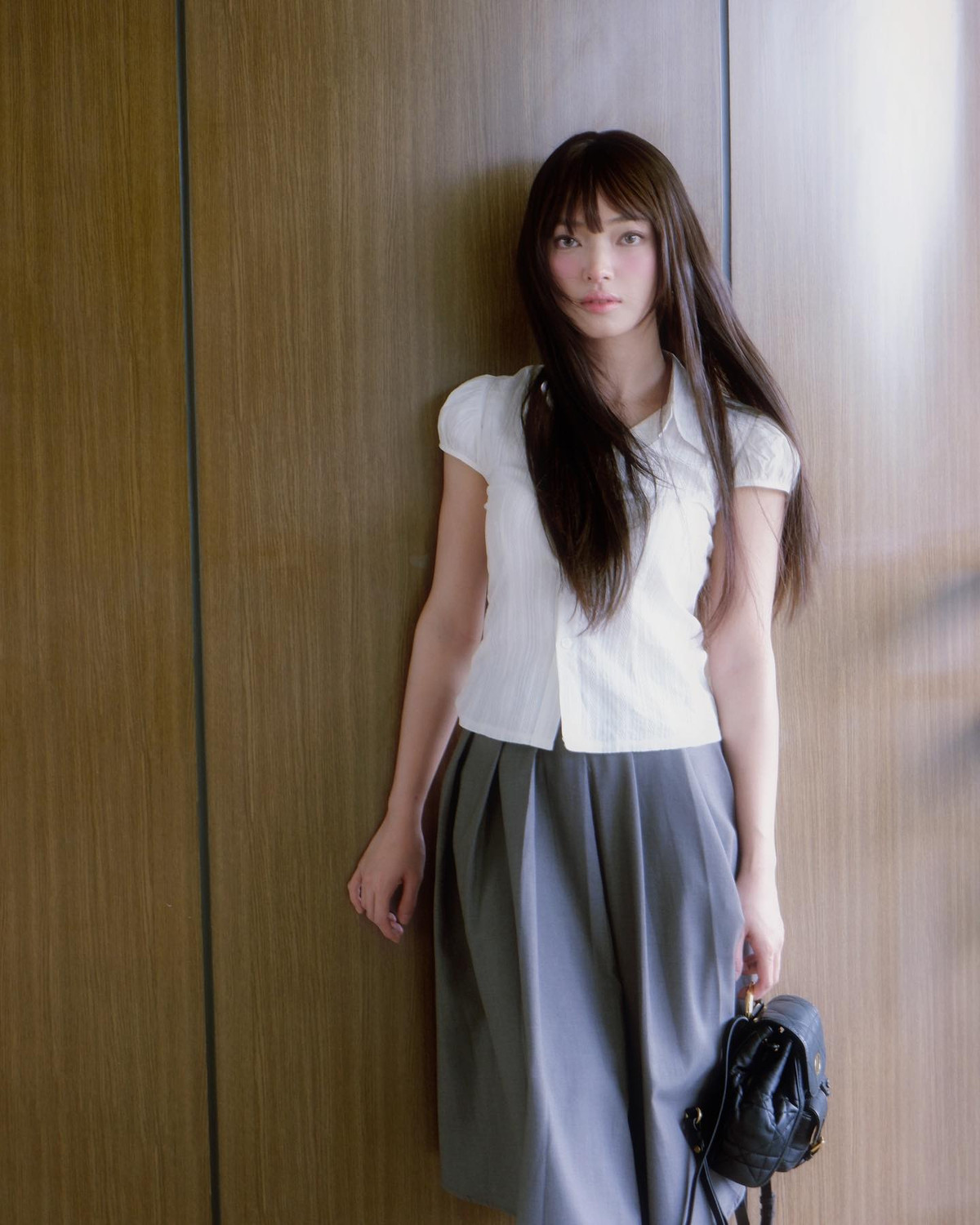 Châu Bùi xuất hiện trong trang phục áo blouse trắng tay bồng dịu dàng, kết hợp với chân váy xếp ly màu ghi dài ngang gối, tạo nên vẻ điệu đà và thanh lịch.