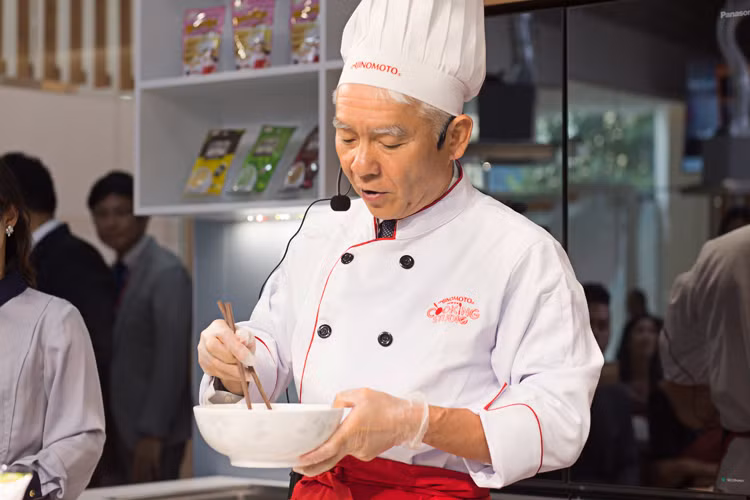 Ông Hiroharu Motohashi bước ra từ khu vực chuẩn bị của nhà bếp Ajinomoto Cooking Studio, khoác lên mình bộ đồ nhà bếp trắng toát, đeo găng tay và hướng dẫn nấu ăn trong sự hoan nghênh, hứng thú của các vị khách.
